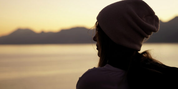 Woman is sitting by the seaside in a cap. Sunset. Cropped photo